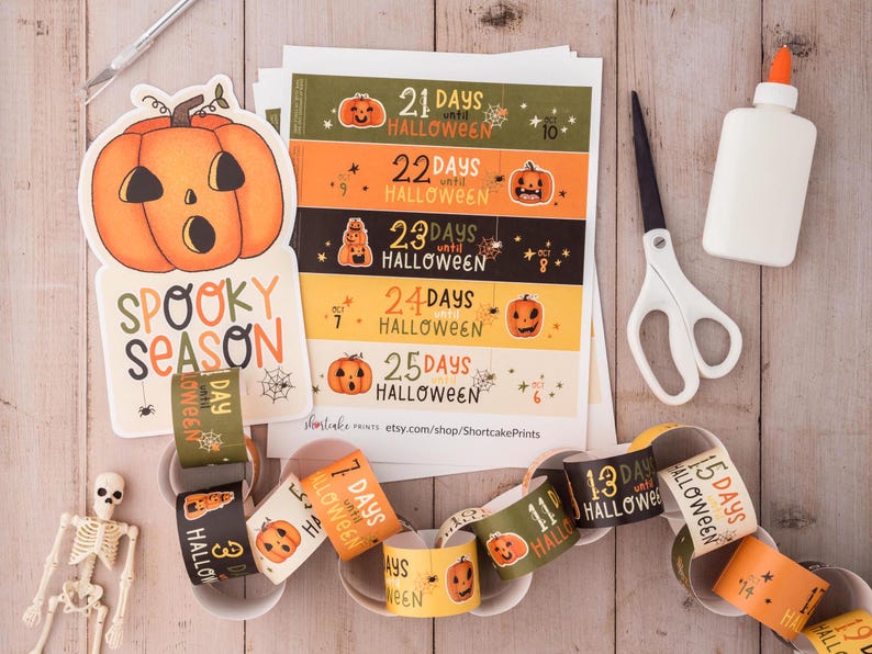 May include: A printable Halloween countdown chain with orange and black paper circles featuring a pumpkin and the text "Days until Halloween". The chain is made of 10 circles and is linked together with glue. The image also includes a printable sheet with 5 different countdown designs, a pair of scissors, a glue bottle, and a large orange pumpkin cutout with the text "Spooky Season".