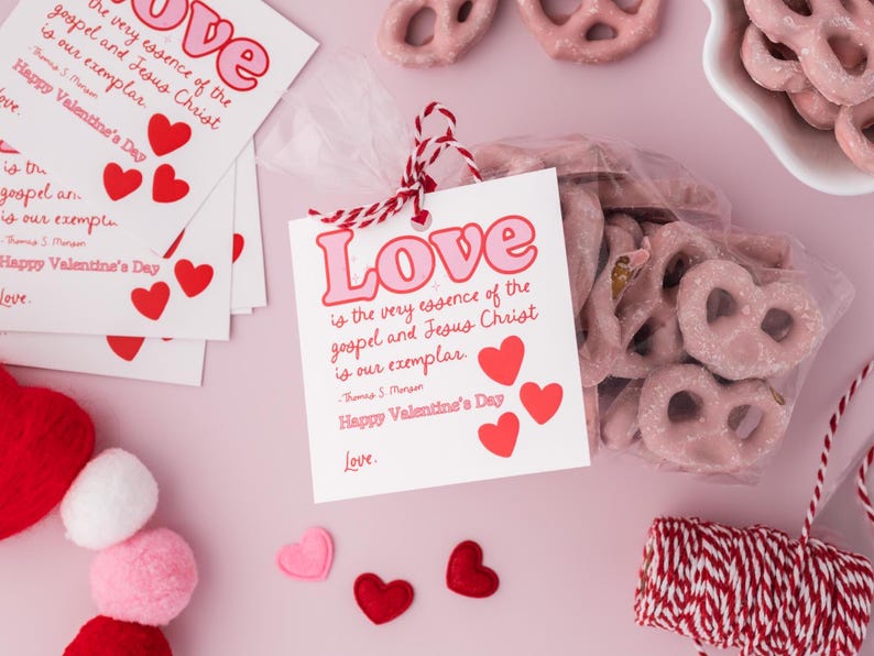K&ouml;nnte beinhalten: Eine rosa und wei&szlig;e Geschenkt&uuml;te gef&uuml;llt mit rosa Brezelbissen und mit roter und wei&szlig;er Schnur gebunden. Eine rosa und wei&szlig;e Karte mit dem Text "Liebe ist die wahre Essenz des Evangeliums und Jesus Christus ist unser Vorbild. Froher Valentinstag. Liebe." ist an der T&uuml;te befestigt.