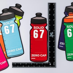 May include: Vibrant water bottle-shaped stickers displaying "SOLID 67" and "ZERO CAP" in white. The stickers are in pink, orange, blue, red, and green. A black ruler, marked in inches, is also visible.