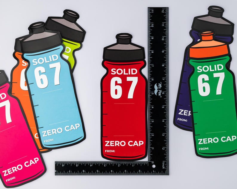 May include: Vibrant water bottle-shaped stickers displaying "SOLID 67" and "ZERO CAP" in white. The stickers are in pink, orange, blue, red, and green. A black ruler, marked in inches, is also visible.