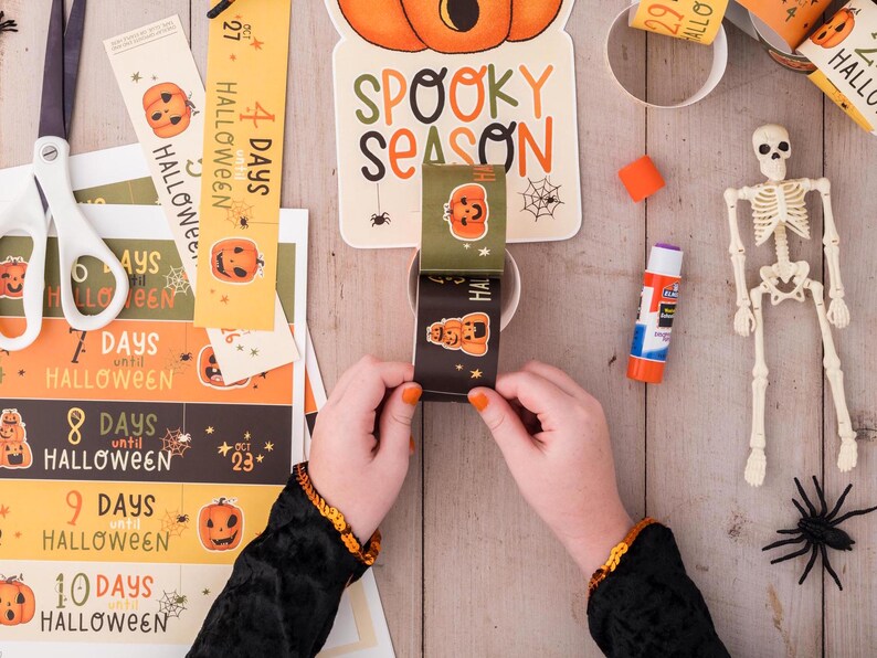 May include: A child's hands are assembling a Halloween countdown with orange and black paper strips featuring pumpkins and the text "Days until Halloween". The strips are being glued to a cardboard tube. There is a skeleton, a spider, and a glue stick on a wooden table.