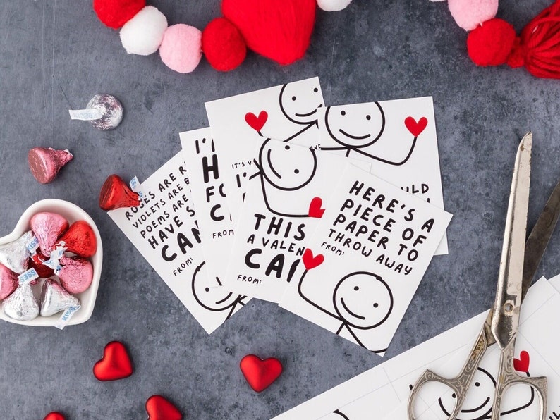 May include: Valentine's Day cards with stick figure illustrations and red heart accents. The cards have humorous messages. The image also includes Hershey's Kisses candies, heart-shaped decorations, and scissors, all arranged on a gray surface.