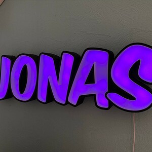 May include: A purple neon sign with the name "JONAS" in a bold, cartoonish font. The sign is made of black plastic with a white backing and has a purple glow.