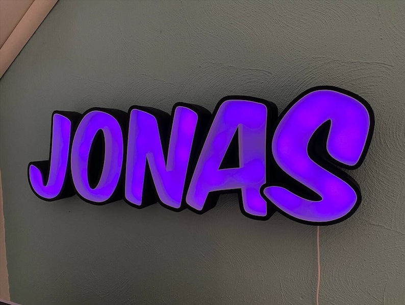 May include: A purple neon sign with the name "JONAS" in a bold, cartoonish font. The sign is made of black plastic with a white backing and has a purple glow.