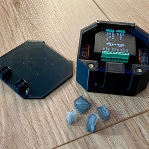 May include: A black 3D printed box with a green circuit board inside. The box is open and the lid is sitting next to it. There are five small gray rubber feet on the table.
