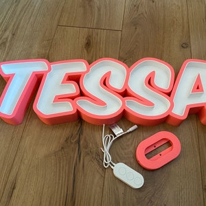 May include: Pink neon sign with the name "TESSA" in white letters. The sign is on a wooden floor and includes a remote control.