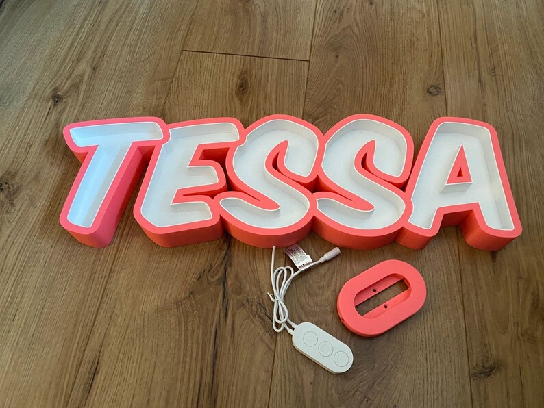 May include: Pink neon sign with the name "TESSA" in white letters. The sign is on a wooden floor and includes a remote control.