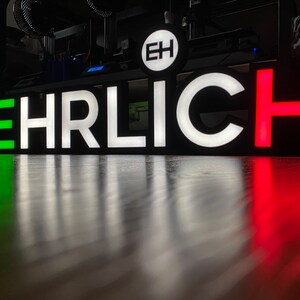 May include: A black sign with the word "EHRLICH" in white letters. The first letter "E" is green, and the last letter "H" is red.