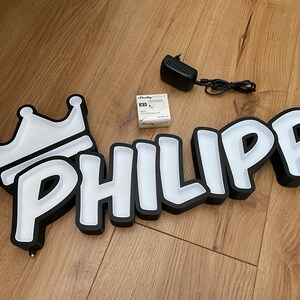 May include: Black and white light-up sign with the name "PHILIPP" in a cartoon font. The sign has a white crown on the left side.