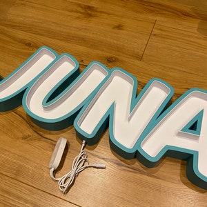 May include: A teal blue light-up name sign spelling "JUNA" with a white interior. The sign is on a wooden surface and has a white cord with a plug.