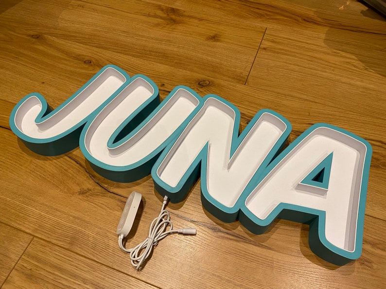 May include: A teal blue light-up name sign spelling "JUNA" with a white interior. The sign is on a wooden surface and has a white cord with a plug.