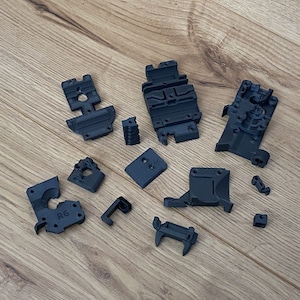 Op de afbeelding: Een set van 13 zwarte 3D-geprinte kunststof onderdelen. De onderdelen zijn in verschillende vormen en maten, waaronder een rechthoekig onderdeel met de tekst "R6" erop gedrukt.