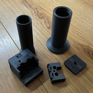 May include: Four black plastic 3D printed parts, including a cylindrical tube, a base with a lip, a rectangular box with the text "R2" on it, and a smaller rectangular piece with a hole in the centre.