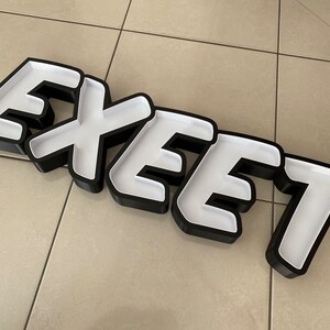 May include: Black and white sign with the word "EXEEET" in bold white letters with a black outline. The sign is made of a thick, rigid material and has a slightly raised, 3D effect.