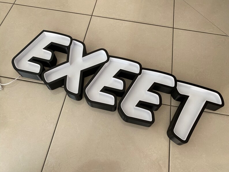 May include: Black and white sign with the word "EXEEET" in bold white letters with a black outline. The sign is made of a thick, rigid material and has a slightly raised, 3D effect.