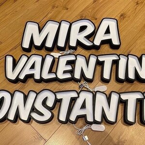 May include: Three white neon signs with black outlines spelling out the names "MIRA", "VALENTIN", and "KONSTANTIN". The signs are on a wooden floor.