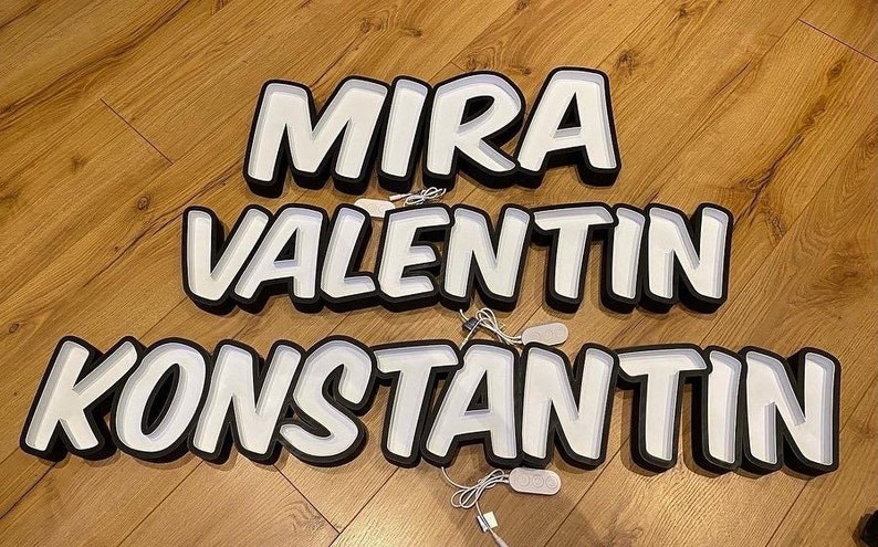 May include: Three white neon signs with black outlines spelling out the names "MIRA", "VALENTIN", and "KONSTANTIN". The signs are on a wooden floor.