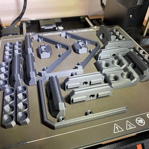 May include: A close-up of a 3D printed model on a printer bed. The model is made of gray plastic and has a variety of shapes and sizes. The printer bed is gray and has a warning label that says "ORIGINAL".