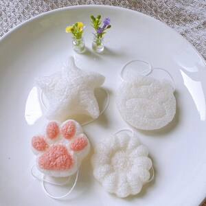 May include: A collection of white bath sponges in various shapes: a star, a cloud, a flower, and a paw print with pink details. The sponges are arranged on a white plate, with small glass vases of flowers in the background.