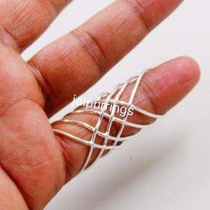 Könnte beinhalten: Ein silberfarbener Fingerring mit einem käfigartigen Design, bestehend aus miteinander verbundenen Stäben, die den Finger umschließen. Der Ring wird auf einem Finger gezeigt, mit dem Text "jaipurrings" sichtbar.
