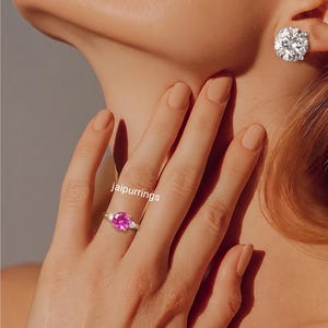 May include: A gold plated ring featuring a round, vibrant pink center stone flanked by two smaller clear stones on each side. The ring is worn on a finger, and a large, round, clear stone stud earring is visible on the earlobe. The text jaipurrings is visible.