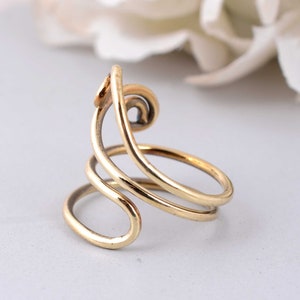May include: A gold-colored ring with a unique, abstract design. The ring features a swirling, openwork pattern with multiple loops and curves. The metal has a polished finish, reflecting light. The ring is set against a soft, neutral background.