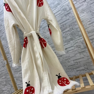 May include: A white cotton robe with a fringed hem and a tie belt. The robe is decorated with red and black ladybug designs.