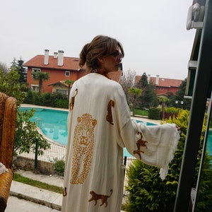 Könnte beinhalten: Cremefarbener Kimono-Robe mit Leopardenmuster. Die Robe hat einen großen Leopardenkopf auf dem Rücken und kleinere Leopardenfiguren. Die Robe hat weite Ärmel und einen Fransenbesatz. Der Hintergrund umfasst einen Pool und Häuser.