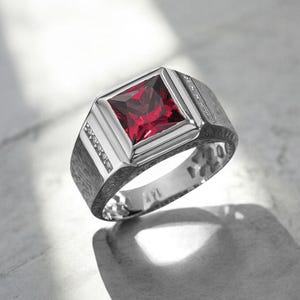 Könnte beinhalten: Silberring mit einem quadratischen, tiefroten Edelstein in der Mitte. Der Ring hat ein strukturiertes Band und kleine Akzentsteine an den Seiten. Der Edelstein ist in einem quadratischen Rahmen gefasst und erzeugt ein kühnes, geometrisches Design.