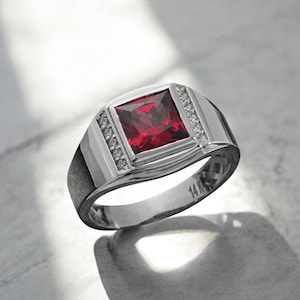 May include: A silver ring with a square, deep red gemstone at the center. The ring is flanked by smaller, clear stones and features a polished finish. The band is marked with "14K".