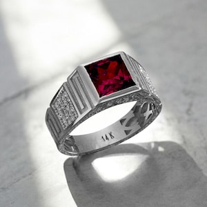 May include: A silver ring with a square, deep red gemstone. The ring features a detailed design with small, clear stones and the inscription "14K". The ring is set on a light-colored surface.