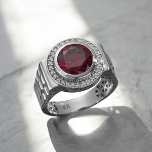 Puede incluir: Un anillo plateado con una gran gema redonda de color rojo intenso en el centro, rodeada por un halo de piedras claras más pequeñas. La banda del anillo tiene un diseño texturizado con cortes geométricos y está estampado con "14K".
