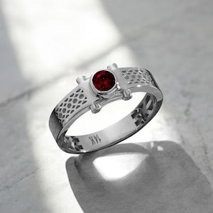 May include: A silver ring with a round, deep red gemstone at the center. The band features a geometric openwork design and small decorative elements on either side of the gemstone. The ring is marked with "14K".