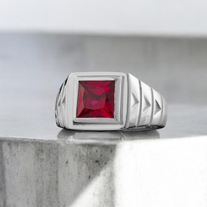 以下が含まれることがあります： 正方形の赤い宝石が付いたシルバーリング。 リングは、隆起したベゼルセッティングを備えた長方形の面を持っています。 バンドは、垂直線と三角形の切り欠きを備えたテクスチャデザインが特徴です。 ライトグレーの表面に表示されています。
