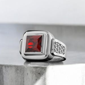 Puede incluir: Un anillo plateado con una gema roja cuadrada y facetada. El anillo tiene una banda gruesa con un patrón geométrico en el lateral. La gema está engastada en un bisel cuadrado.