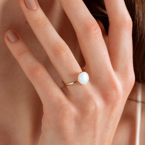 Puede incluir: Un anillo de oro con una piedra de ópalo blanco. El anillo está en el dedo de una persona.