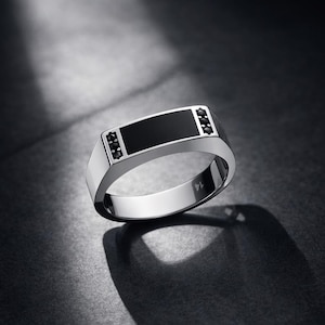 May include: A polished silver ring featuring a rectangular black onyx centre stone, flanked by three small black stones on each side. The ring's design is modern, set against a dark backdrop, emphasizing its sleek, geometric form.