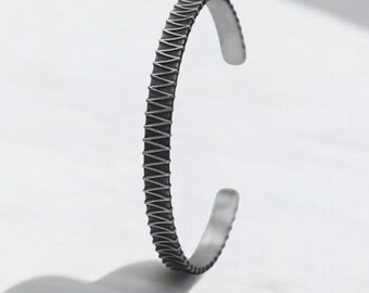 May include: A silver-colored cuff bracelet with a textured, zig-zag pattern. The open-ended design allows for easy wear. The bracelet is displayed on a white surface, with a shadow cast to the left.