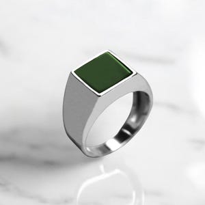 Op de afbeelding: Een zilveren ring met een vierkante groene steen in het midden.