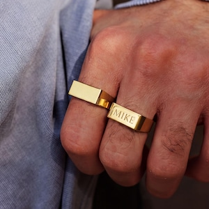 Könnte beinhalten: Zwei goldfarbene, rechteckige Siegelringe an nebeneinanderliegenden Fingern. Ein Ring hat eine glatte, leere Oberfläche, während der andere Ring mit dem Namen MIKE in Großbuchstaben graviert ist. Beide Ringe haben eine polierte Oberfläche.