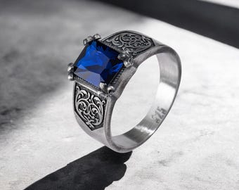 May include: A silver ring with a square, deep blue gemstone. The ring features ornate scrollwork on the band and around the gemstone setting. The ring is set on a light gray surface, with a shadow cast to the side.
