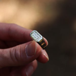 以下が含まれることがあります： 1997年がブロック体で刻印された長方形の面を持つ、シルバーカラーのシグネットリング。リングは光沢のある金属仕上げで、上面には面取りが施されており、滑らかで先細りのバンドデザインが特徴です。