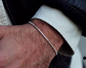 Pode incluir: Uma pulseira de corrente fina tipo caixa, de cor prateada com um acabamento metálico polido. A pulseira apresenta um design contínuo de elos quadrados que envolve o pulso, exibindo um padrão uniforme de pequenos segmentos metálicos refletores.