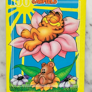 Puede incluir: Un rompecabezas de 100 piezas con Garfield el gato, acostado sobre una flor rosa, con un osito de peluche sentado sobre una flor amarilla en primer plano. La caja del rompecabezas es amarilla con un fondo azul y las palabras "100 GARFIELD" en blanco.