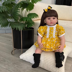 May include: A doll with brown hair and bangs, wearing a yellow dress with a white daisy pattern, white lace trim, and a matching yellow and black hair bow. The doll has black socks with black bows and is seated on a white textured pillow.