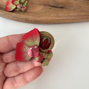 Könnte beinhalten: Eine handgefertigte Brosche aus Polymer Clay mit einem Erdbeer- und Herzdesign in Rot und Grün. Das Herz hat eine glitzernde Oberfläche.