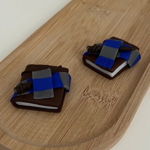 Può includere: Due piccoli libri marroni con una sciarpa blu e grigia e una bacchetta marrone in cima. I libri sono su una superficie di legno.