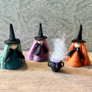 May include: Three small witch figurines with black felt hats and yarn hair in teal, purple, and orange. Each wears a black cape over a matching dress. A miniature black cauldron sits in the centre, filled with purple liquid and white fibre smoke.
