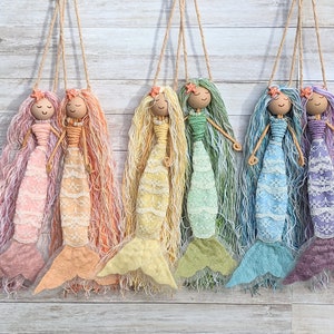 May include: Six colourful mermaid dolls with long yarn hair and lace dresses hanging from strings against a wood background. The mermaids are pink, orange, yellow, green, blue, and purple.