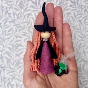 May include: A small witch figurine with a black felt hat, long peach-coloured yarn hair, and a purple felt dress, featuring a wooden bead head and a tiny black cauldron with green liquid and white wispy smoke accents attached to the side.